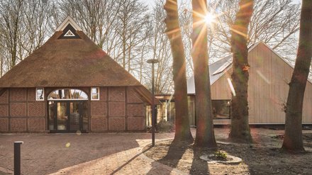 Das Foto zeigt ein Ensemble aus zwei Gebäuden: links ein traditionelles Fachwerkhaus mit Reetdach und bogenförmiger Glastür, rechts ein modernes Haus mit Holzfassade. Beide sind durch einen transparenten Gang verbunden und liegen in einem von Bäumen umgebenen Gelände. Ein gepflasterter Weg führt auf sie zu, die Sonne wirft lange Schatten.