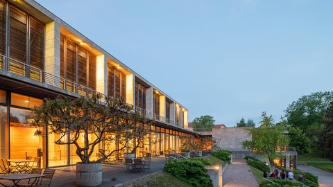 Modernes Gebäude mit beleuchteten großen Glasfenstern und Holzjalousien  in der Abenddämmerung. Terrassierte Außenfläche mit Bäumen in Kübeln  und Sitzmöbeln, Besucher sitzen auf einer Wiese.