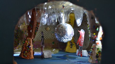 Blick durch einen Tunnel auf eine bunte, mit Glitzer verzierte Innenraumszene mit einer silbernen Kugel in der Mitte und kleinen Figuren aus Papier und Pappe