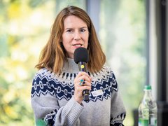 „Eine Frau mit braunen Haaren und gemustertem Pullover spricht bei einer Veranstaltung in ein Handmikrofon.“