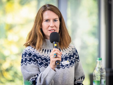 „Eine Frau mit braunen Haaren und gemustertem Pullover spricht bei einer Veranstaltung in ein Handmikrofon.“
