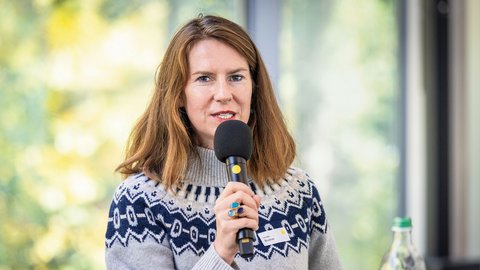 „Eine Frau mit braunen Haaren und gemustertem Pullover spricht bei einer Veranstaltung in ein Handmikrofon.“
