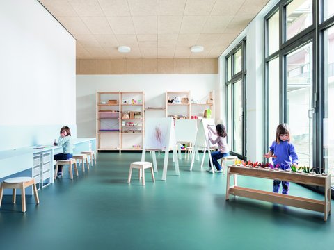 "Heller Kreativraum mit großem Fensterfront. Links Arbeitstische mit Stühlen, dahinter offene Holzregale mit Materialien und Boxen. Mitte: mehrere Staffeleien zum Malen mit Kindern. Rechts ein Arbeitstisch mit Stiften und Materialien. Fünf Kinder in verschiedenen Farben gekleidet arbeiten an verschiedenen Stationen. Beige Decke mit Beleuchtung, türkiser Bodenbelag."