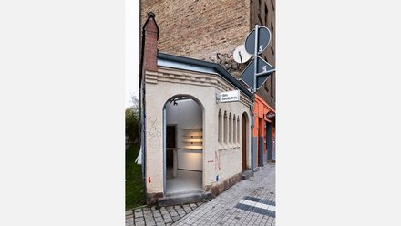 Kleines historisches Eckgebäude „das Bedürfnis“ zwischen hohen Häusern; renovierter heller Putz und offener Eingang zeigen den Ausstellungsraum im Inneren