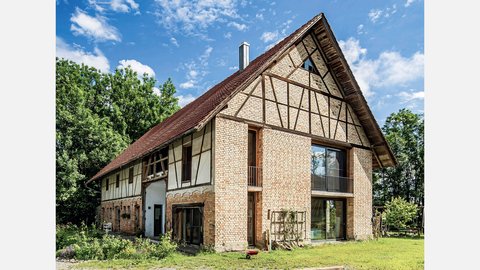 Außenansicht eines großen, zweistöckigen Wohnhauses mit historischem Fachwerk-Design. Das Gebäude kombiniert beige-rote Backsteine mit dunkelbraunen Holzbalken in charakteristischem Fachwerk-Muster. Das Dach ist mit roten Dachziegeln gedeckt und hat einen ausgeprägten Giebel. Große moderne Fenster und Glastüren sind in die historische Struktur integriert. Das Haus ist von grüner Vegetation umgeben, mit Büschen und Bäumen im Hintergrund.
