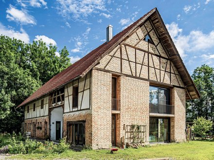 Außenansicht eines großen, zweistöckigen Wohnhauses mit historischem Fachwerk-Design. Das Gebäude kombiniert beige-rote Backsteine mit dunkelbraunen Holzbalken in charakteristischem Fachwerk-Muster. Das Dach ist mit roten Dachziegeln gedeckt und hat einen ausgeprägten Giebel. Große moderne Fenster und Glastüren sind in die historische Struktur integriert. Das Haus ist von grüner Vegetation umgeben, mit Büschen und Bäumen im Hintergrund.