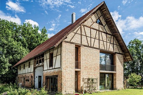 Außenansicht eines großen, zweistöckigen Wohnhauses mit historischem Fachwerk-Design. Das Gebäude kombiniert beige-rote Backsteine mit dunkelbraunen Holzbalken in charakteristischem Fachwerk-Muster. Das Dach ist mit roten Dachziegeln gedeckt und hat einen ausgeprägten Giebel. Große moderne Fenster und Glastüren sind in die historische Struktur integriert. Das Haus ist von grüner Vegetation umgeben, mit Büschen und Bäumen im Hintergrund.