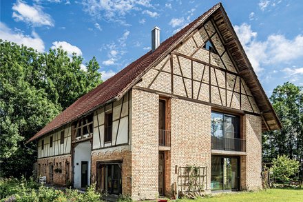 Außenansicht eines großen, zweistöckigen Wohnhauses mit historischem Fachwerk-Design. Das Gebäude kombiniert beige-rote Backsteine mit dunkelbraunen Holzbalken in charakteristischem Fachwerk-Muster. Das Dach ist mit roten Dachziegeln gedeckt und hat einen ausgeprägten Giebel. Große moderne Fenster und Glastüren sind in die historische Struktur integriert. Das Haus ist von grüner Vegetation umgeben, mit Büschen und Bäumen im Hintergrund.