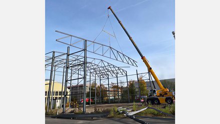 Das Bild zeigt den Aufbau einer großen Stahlkonstruktion im Freien. Ein gelber Mobilkran hebt ein großes Stahlträgerelement in die Höhe, während bereits montierte Stahlstützen und Dachträger ein offenes Hallengerüst bilden. Der Himmel ist klar und blau, im Hintergrund stehen weitere Gebäude. Die Stahlkonstruktion wirkt industriell und modular. Zweck des Bildes ist es, den Montageprozess wiederverwendbarer Stahlbauteile zu dokumentieren und zu zeigen, wie bestehende Konstruktionen erneut aufgebaut und in neue Bauprojekte integriert werden können.