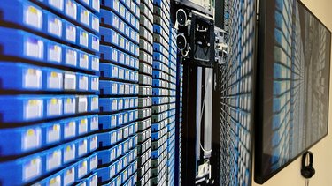 Ein großer Bildschirm zeigt eine extreme Nahaufnahme von dicht gestapelten Datenträgern oder Kassetten in Blau- und Schwarztönen. Daneben hängt ein Kopfhörer. Das Bild verweist auf digitale Speicherung und Archivierung. Der Kopfhörer deutet darauf hin, dass es sich um eine audiovisuelle Arbeit handelt, die zusätzlich hörbare Informationen vermittelt.