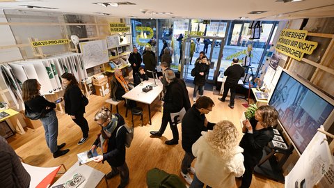 Besucherinnen und Besucher bewegen sich durch einen Ausstellungsraum mit Infomaterial und Tischen.