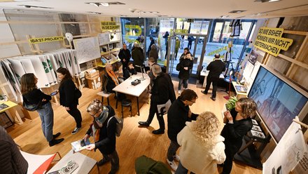 Besucherinnen und Besucher bewegen sich durch einen Ausstellungsraum mit Infomaterial und Tischen.