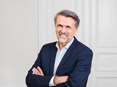 Porträtfoto eines mittelalten Mannes mit grauem Haar und Bartansatz. Er trägt einen dunkelblauen Blazer über einem hellblauen Hemd. Die Arme sind verschränkt vor der Brust. Er lächelt selbstbewusst in die Kamera. Der Hintergrund zeigt eine neutrale weiße Wand mit vertikalen Paneelen. Professionelle Studio-Beleuchtung."