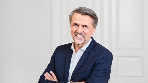 Porträtfoto eines mittelalten Mannes mit grauem Haar und Bartansatz. Er trägt einen dunkelblauen Blazer über einem hellblauen Hemd. Die Arme sind verschränkt vor der Brust. Er lächelt selbstbewusst in die Kamera. Der Hintergrund zeigt eine neutrale weiße Wand mit vertikalen Paneelen. Professionelle Studio-Beleuchtung."