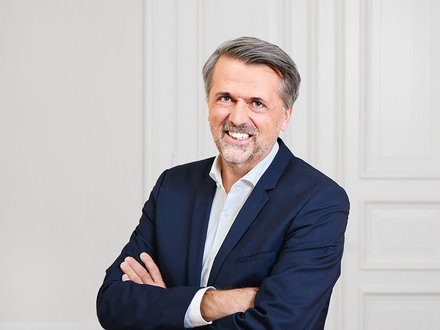 Porträtfoto eines mittelalten Mannes mit grauem Haar und Bartansatz. Er trägt einen dunkelblauen Blazer über einem hellblauen Hemd. Die Arme sind verschränkt vor der Brust. Er lächelt selbstbewusst in die Kamera. Der Hintergrund zeigt eine neutrale weiße Wand mit vertikalen Paneelen. Professionelle Studio-Beleuchtung."