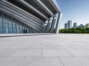 Blick über eine große, gepflasterte Fläche auf ein modernes Gebäude mit schrägen Stützen und Glasfassade, im Hintergrund Stadtgebäude und Bäume.