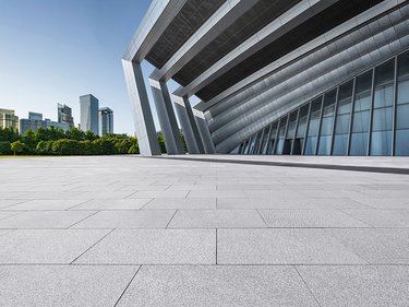 Blick über eine große, gepflasterte Fläche auf ein modernes Gebäude mit schrägen Stützen und Glasfassade, im Hintergrund Stadtgebäude und Bäume.