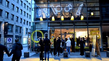 Abendliche Außenansicht eines Glasgebäudes mit beleuchtetem Innenraum und wartenden Menschen.