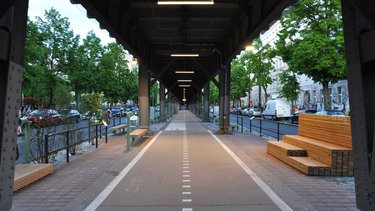 Zentralperspektive des Radwegs unter der Hochbahn, beleuchtete Konstruktion, Sitzgelegenheiten und bepflanzte Randbereiche.