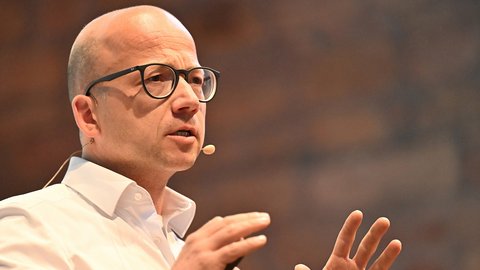 "Nahaufnahme eines älteren Mannes im Profil mit kahlem Kopf, schwarzer Brille und weißem Hemd mit beiger Krawatte. Er spricht engagiert und gestikuliert mit beiden Händen. Ein Headset-Mikrofon ist sichtbar. Der unscharfe Hintergrund zeigt warme braune Farbtöne."