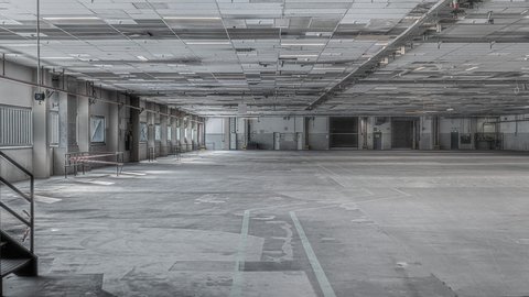 Leere, weitläufige Industriehalle: grauer Betonboden mit verblassten Markierungen, niedrige Rasterdecke mit Leitungen und Lichtbändern; links Fensterreihe und Treppe, hinten Laderampen und Tore.