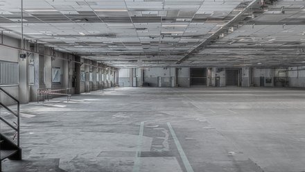 Leere, weitläufige Industriehalle: grauer Betonboden mit verblassten Markierungen, niedrige Rasterdecke mit Leitungen und Lichtbändern; links Fensterreihe und Treppe, hinten Laderampen und Tore.