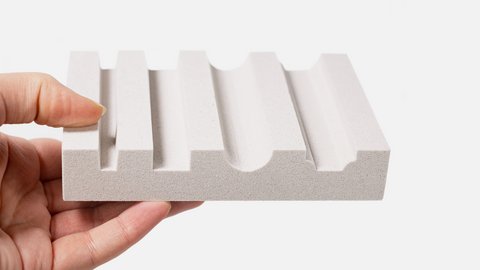 Hand hält eine hellgraue, feinporige Materialprobe mit mehreren parallel verlaufenden Rillen – abwechselnd rechteckig und halbkreisförmig gefräst.