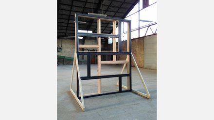 Freistehende Wandprobe aus mehreren rechteckigen Holz‑ und Aluminium‑Fensterrahmen in gestaffelter Anordnung, abgestützt auf diagonalen Holzstreben; Aufnahme in einer großen, hellen Industriehalle mit sichtbarer Dachkonstruktion und Backsteinwand im Hintergrund.