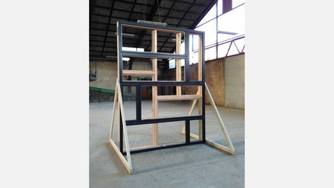 Freistehende Wandprobe aus mehreren rechteckigen Holz‑ und Aluminium‑Fensterrahmen in gestaffelter Anordnung, abgestützt auf diagonalen Holzstreben; Aufnahme in einer großen, hellen Industriehalle mit sichtbarer Dachkonstruktion und Backsteinwand im Hintergrund.