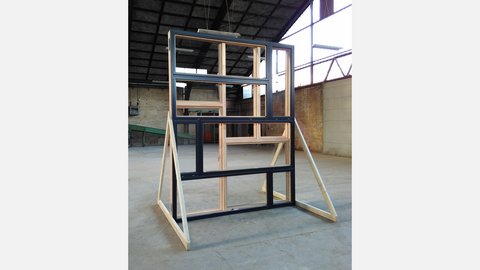 Freistehende Wandprobe aus mehreren rechteckigen Holz‑ und Aluminium‑Fensterrahmen in gestaffelter Anordnung, abgestützt auf diagonalen Holzstreben; Aufnahme in einer großen, hellen Industriehalle mit sichtbarer Dachkonstruktion und Backsteinwand im Hintergrund.
