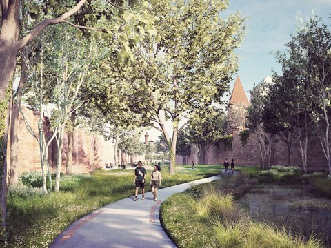 Schmaler gepflasterter Fußweg mit rotem Ziegelbelag entlang eines flachen Wasserlaufs. Hohe Bäume säumen beide Seiten. Paare joggen/gehen auf dem Weg. Historische Steinmauern und Gebäudetürme sichtbar. Grüne Vegetation und Wiese.
