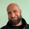 "Portraitfoto eines Mannes mittleren Alters mit Glatze und vollem, dunklem Bart. Er trägt ein schwarzes Langarmshirt mit einer schwarzen Fleecejacke mit Reißverschluss. Im Hintergrund eine hellgrüne Wand. Der Mann blickt leicht zur Seite und lächelt freundlich."
