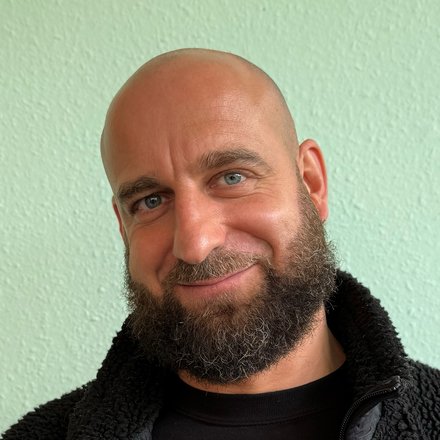 "Portraitfoto eines Mannes mittleren Alters mit Glatze und vollem, dunklem Bart. Er trägt ein schwarzes Langarmshirt mit einer schwarzen Fleecejacke mit Reißverschluss. Im Hintergrund eine hellgrüne Wand. Der Mann blickt leicht zur Seite und lächelt freundlich."