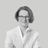 Schwarzweiß-Porträt einer lächelnden Frau mit kurzer Frisur und Brille, frontal aufgenommen, sie trägt ein helles Sakko und ein Hemd, hat die Arme verschränkt; neutraler, einfarbiger Hintergrund.