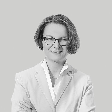 Schwarzweiß-Porträt einer lächelnden Frau mit kurzer Frisur und Brille, frontal aufgenommen, sie trägt ein helles Sakko und ein Hemd, hat die Arme verschränkt; neutraler, einfarbiger Hintergrund.