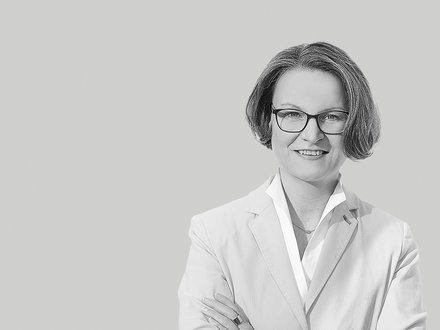 Schwarzweiß-Porträt einer lächelnden Frau mit kurzer Frisur und Brille, frontal aufgenommen, sie trägt ein helles Sakko und ein Hemd, hat die Arme verschränkt; neutraler, einfarbiger Hintergrund.