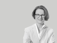 Schwarzweiß-Porträt einer lächelnden Frau mit kurzer Frisur und Brille, frontal aufgenommen, sie trägt ein helles Sakko und ein Hemd, hat die Arme verschränkt; neutraler, einfarbiger Hintergrund.
