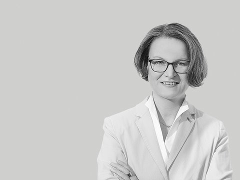 Schwarzweiß-Porträt einer lächelnden Frau mit kurzer Frisur und Brille, frontal aufgenommen, sie trägt ein helles Sakko und ein Hemd, hat die Arme verschränkt; neutraler, einfarbiger Hintergrund.