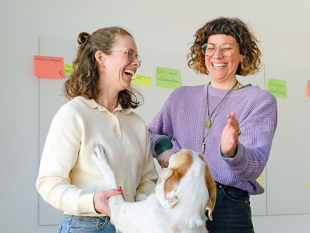Zwei lachende Frauen stehen vor einer hellgrauen Wand mit farbigen Haft-Notizen in Rot, Orange und Grün. Die Frau links trägt ein cremefarbenes Sweatshirt und Jeans, die Frau rechts trägt einen violetten Pullover und schwarze Hose. Zwischen ihnen springt ein kleiner weiß-braun gefleckter Hund mit längeren Haaren. Beide Frauen tragen Brillen und wirken freudig. Im Hintergrund Notizen auf der Pinnwand