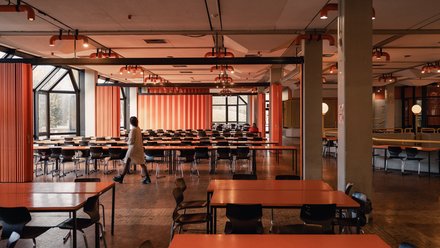 Großer Speisesaal bei Tageslicht: orange Tische, schwarze Stühle, orange Deckenleuchten und gestreifte Faltwände; links Fensterfront, eine Person geht durch den Mittelgang.