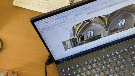 Tisch mit einem Laptop auf dessen Bildschirm ein Haus zu sehen ist. Notizheft im Hintergrund des Tisches 