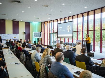 Großer Konferenzraum mit vielen sitzenden Teilnehmenden, vorne hält eine Person am Pult einen Vortrag