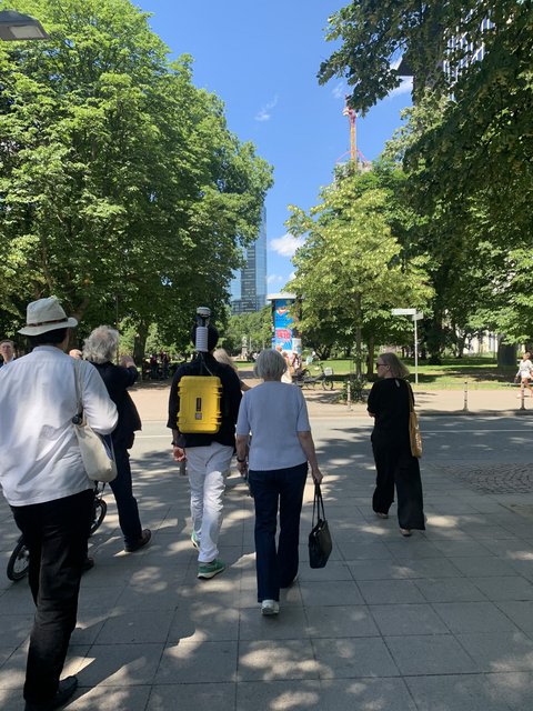 Eine kleine Gruppe von Menschen spaziert auf einem Gehweg an einem Park entlang, vor ihnen läuft jemand mit einem gelben Koffer-Rucksack.