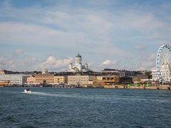 "Stadtansicht einer nordischen Hafenstadt von der Wasserseite aus. Im Vordergrund ein Motorboot auf Wasser. Die Skyline zeigt historische gelbliche und orange Gebäude verschiedener Größen, eine markante Kathedrale mit grüner Kuppel in der Mitte und ein großes weißes Riesenrad mit blauen Gondeln auf der rechten Seite. Ein blauer Himmel mit weißen Wolken und Baustellenkräne sind oben sichtbar."