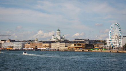 "Stadtansicht einer nordischen Hafenstadt von der Wasserseite aus. Im Vordergrund ein Motorboot auf Wasser. Die Skyline zeigt historische gelbliche und orange Gebäude verschiedener Größen, eine markante Kathedrale mit grüner Kuppel in der Mitte und ein großes weißes Riesenrad mit blauen Gondeln auf der rechten Seite. Ein blauer Himmel mit weißen Wolken und Baustellenkräne sind oben sichtbar."