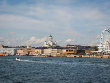 "Stadtansicht einer nordischen Hafenstadt von der Wasserseite aus. Im Vordergrund ein Motorboot auf Wasser. Die Skyline zeigt historische gelbliche und orange Gebäude verschiedener Größen, eine markante Kathedrale mit grüner Kuppel in der Mitte und ein großes weißes Riesenrad mit blauen Gondeln auf der rechten Seite. Ein blauer Himmel mit weißen Wolken und Baustellenkräne sind oben sichtbar."