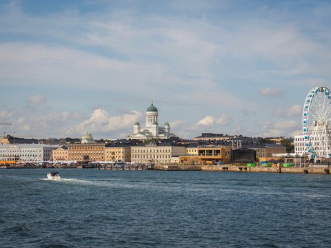 "Stadtansicht einer nordischen Hafenstadt von der Wasserseite aus. Im Vordergrund ein Motorboot auf Wasser. Die Skyline zeigt historische gelbliche und orange Gebäude verschiedener Größen, eine markante Kathedrale mit grüner Kuppel in der Mitte und ein großes weißes Riesenrad mit blauen Gondeln auf der rechten Seite. Ein blauer Himmel mit weißen Wolken und Baustellenkräne sind oben sichtbar."
