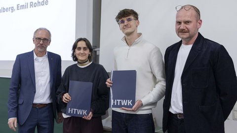Vier Personen vor einer Leinwand; zwei halten Mappen mit Urkunden – Moment einer Preisverleihung.