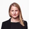 "Porträtfoto einer Frau mit schulterlangen, hellbraunen Haaren. Sie trägt einen schwarzen Blazer über einem schwarzen Shirt und blickt selbstbewusst in die Kamera. Die Hände befinden sich in den Hosentaschen. Der Hintergrund ist weiß."