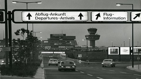 Eine Schwarz-Weiß-Fotografie bei Dämmerung oder Nacht zeigt die Zufahrt zum Flughafen Berlin-Tegel. Im Vordergrund ist eine Straße mit mehreren Autos zu sehen. Ein großes, beleuchtetes Hinweisschild über der Straße zeigt Pfeile und die Aufschrift "Abflug-Ankunft Departures-Arrivals" sowie "Flug-Information". Im Hintergrund sind das beleuchtete Terminalgebäude mit dem Schriftzug "BERLIN-TEGEL" und der Kontrollturm zu erkennen. Die Szene wird von Straßenlaternen und Lichtern der Gebäude erhellt und wirkt geschäftig.
